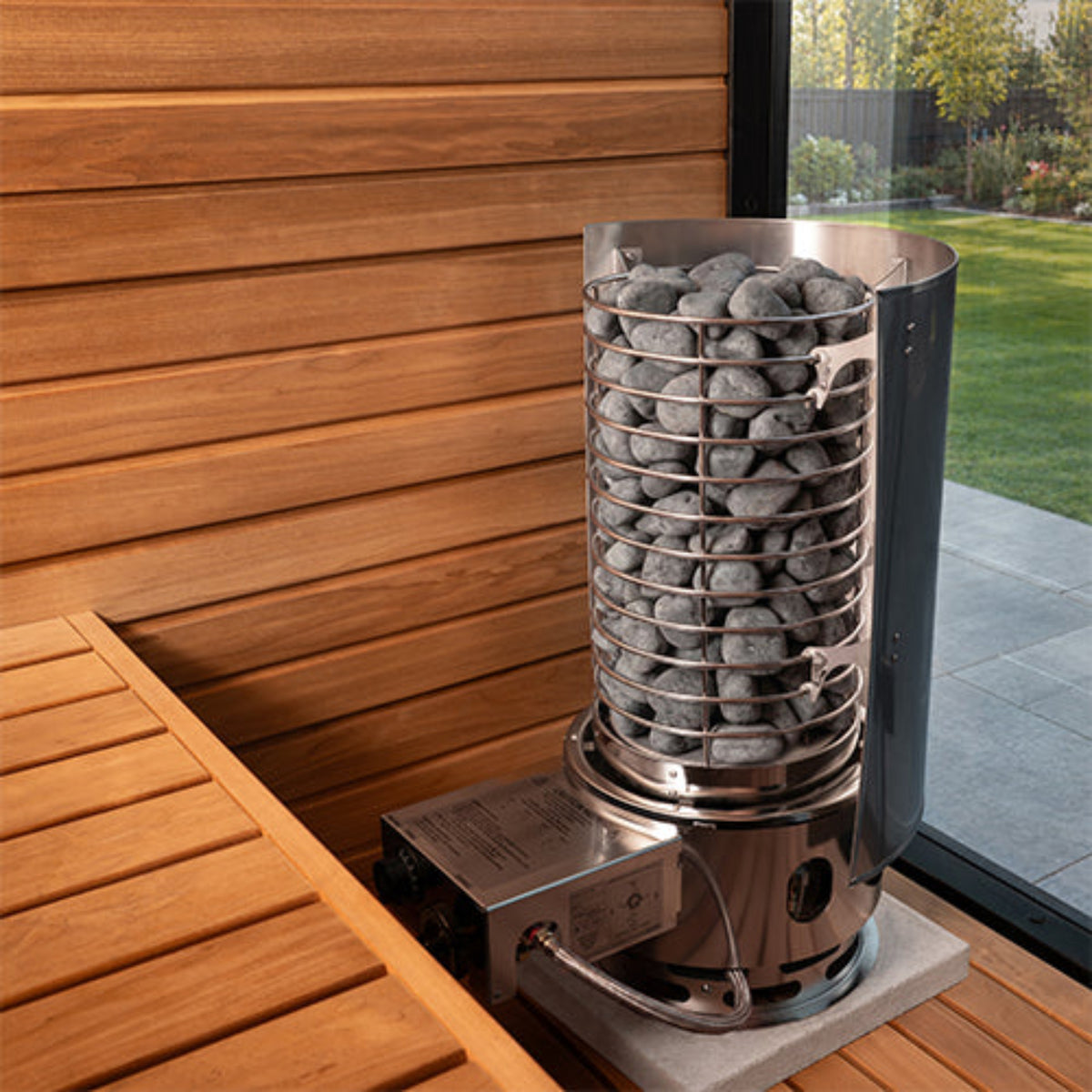 A stainless steel stone heater with gray heating stones inside a wire cage, shown on a wooden deck next to a window with a garden view.
