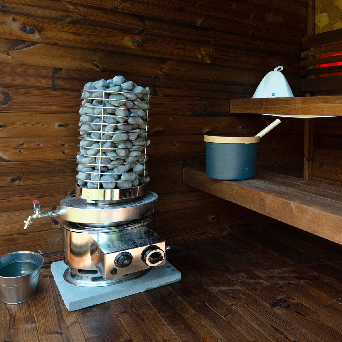 A stainless steel sauna stone heater filled with gray stones, set on a wooden floor inside a wooden room with a blue bucket and wooden shelves.