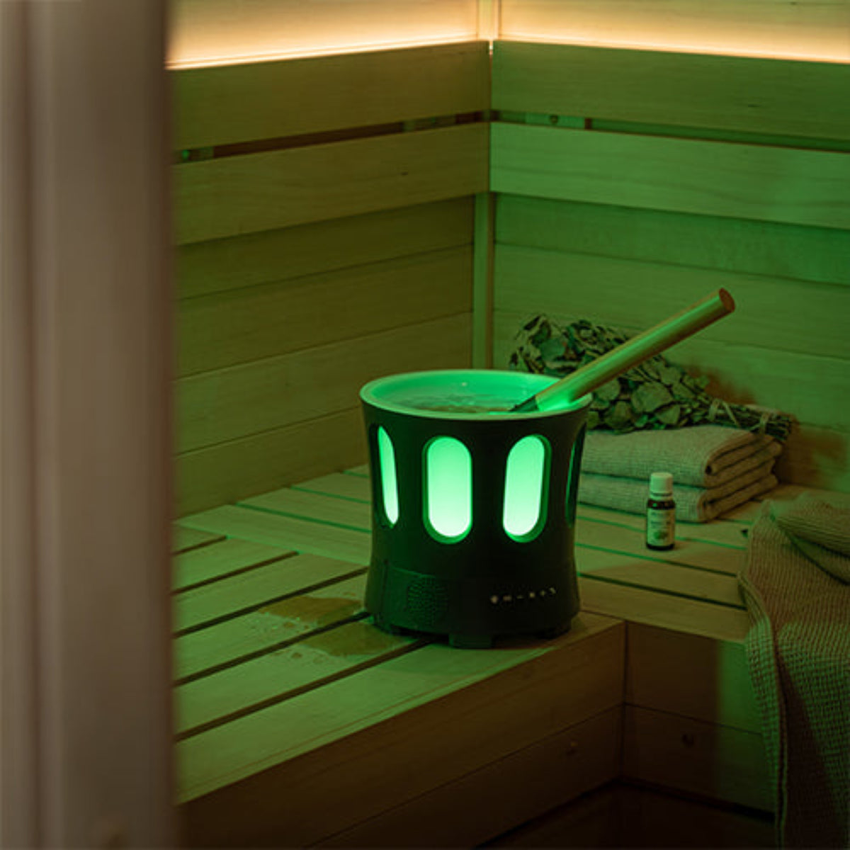 Bluetooth Sauna Bucket & Bamboo Ladle with glowing green LED light panels, placed on a wooden sauna bench with a bamboo scoop and a small bottle nearby.
