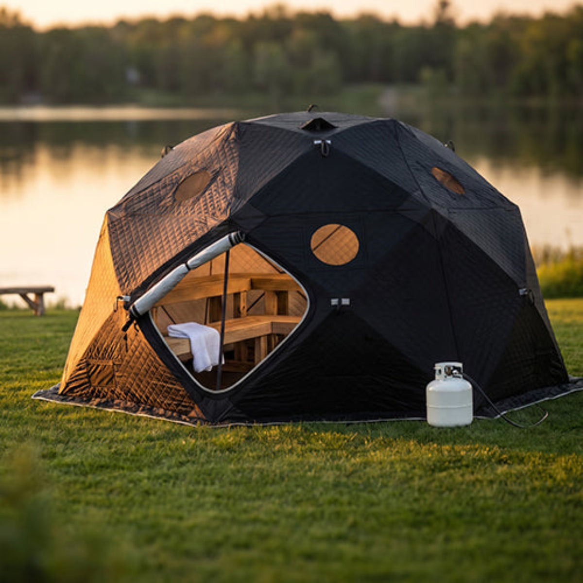 SaunaLife SaunaTrekker XL Portable Sauna Tent pitched on a grassy lawn near a lake at sunset, with a small white propane tank beside it and trees in the background.