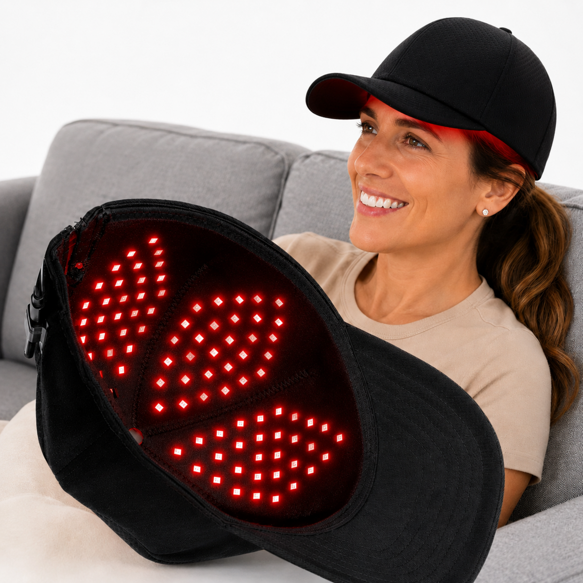 A woman wearing a black Red Light Pro Device cap sits on a gray couch holding the cap open to show a red light therapy panel inside. The cap is shown both worn and opened to display the illuminated interior.