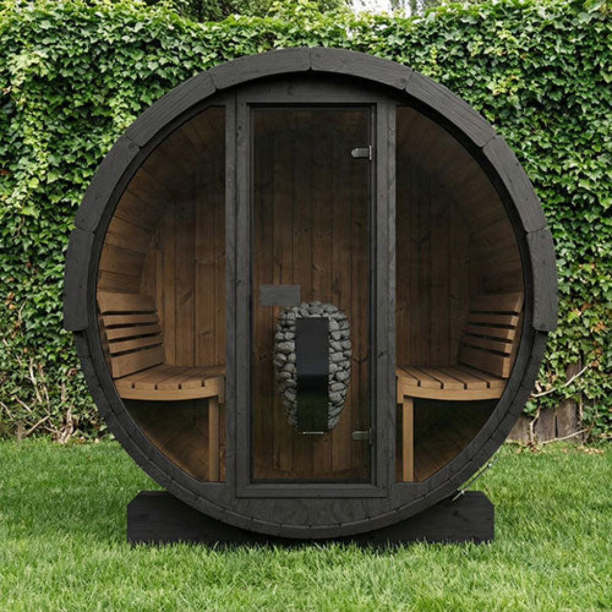 SaunaLife ERGO-Series Model E7G Outdoor Barrel Sauna with a dark charcoal exterior and warm brown wood interior, featuring two built-in benches and a glass door with a stone fire insert, set outdoors on grass in front of a leafy hedge.
