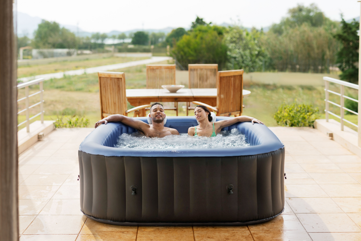 Two people in an inflatable hot tub on a patio with a scenic background