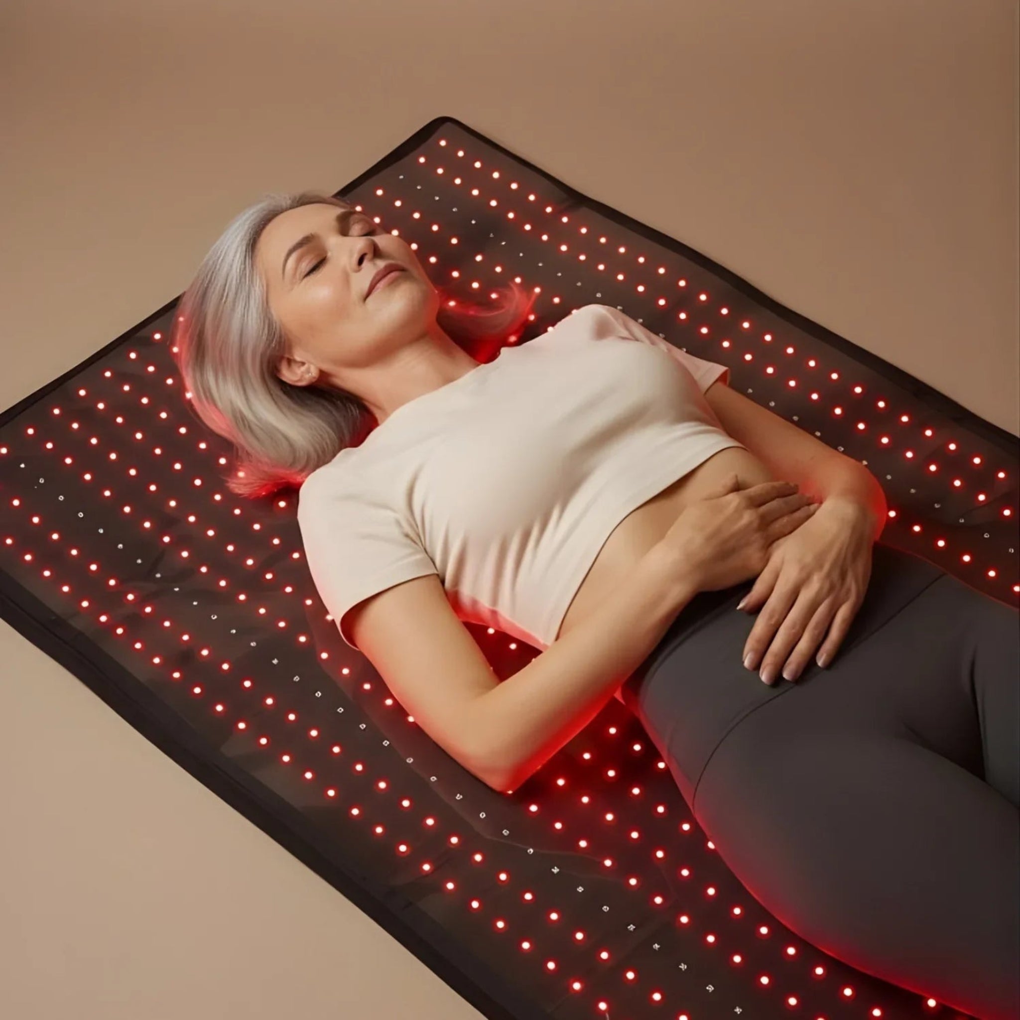 A woman lies on a black LED light therapy mat with glowing red lights, against a warm beige background.