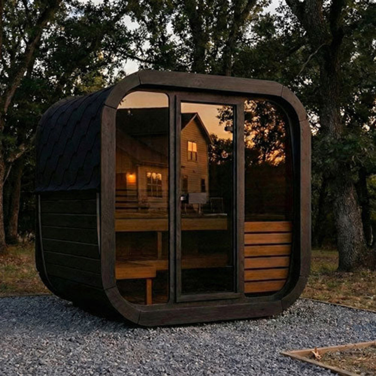 SaunaLife CUBE-Series Model CL5GB Outdoor Cube Sauna, shown on a gravel surface in a wooded setting at dusk. The cabin reflects a house and trees, and warm light is visible inside.
