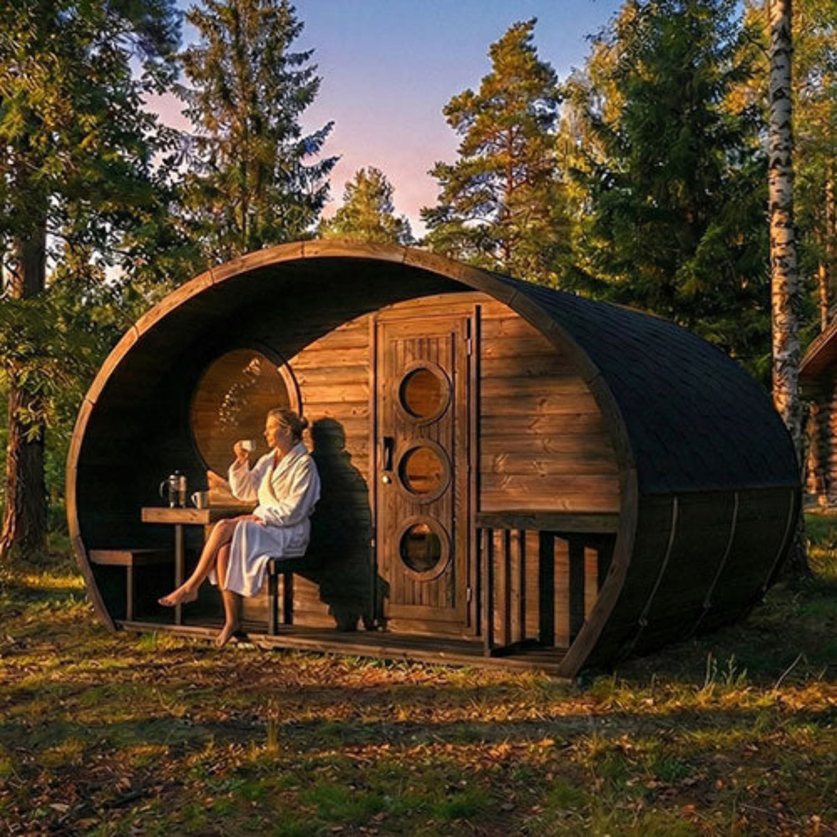 SaunaLife G11B outdoor garden sauna suite sits in a forest clearing at sunset, with a woman sitting inside near a small table. Tall evergreen trees surround the cabin, and the ground is grassy with fallen leaves.