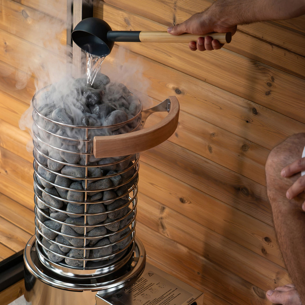 A person pours hot water into a metal stone sauna steamer filled with dark heating stones, set against a warm wood-paneled wall.