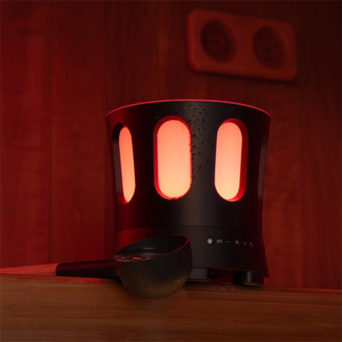 Bluetooth Sauna Bucket & Bamboo Ladle with three vertical oval cutouts glowing warm orange-red light, placed on a wooden surface against a dark wood wall. 