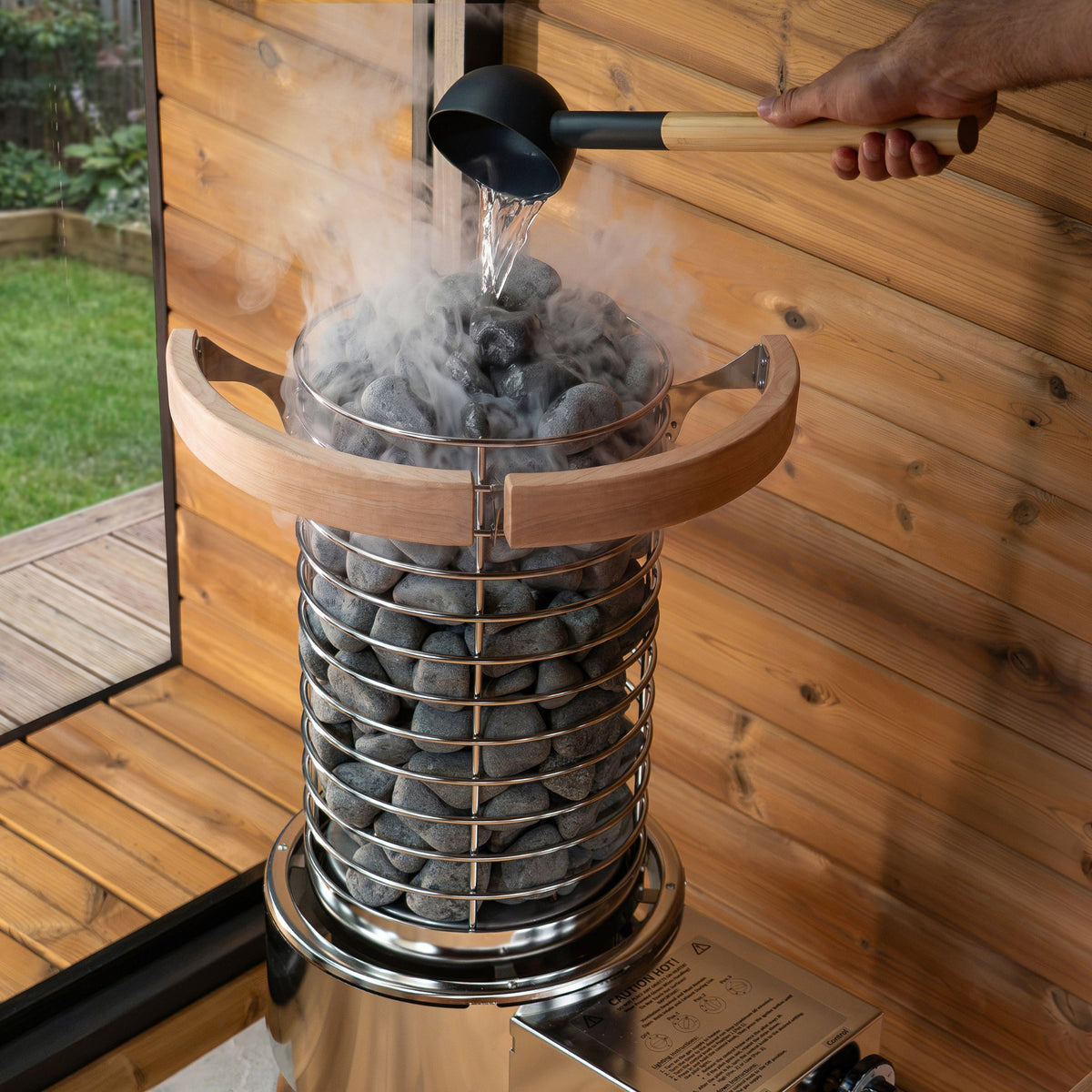 A sauna stone boiler with a metal mesh basket filled with dark heating stones, shown with steam rising as water is being poured into it from a ladle. The unit is set against a wooden wall and deck background.
