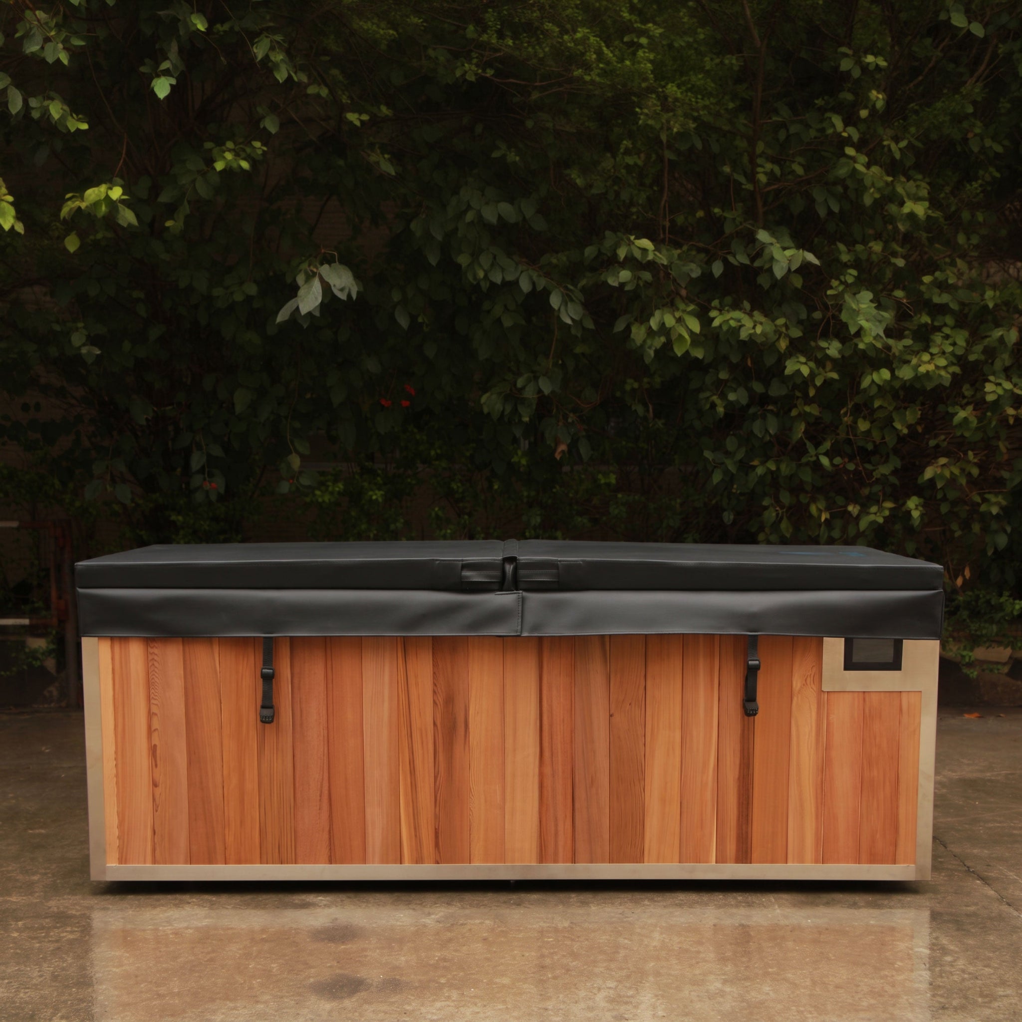 Finnmark plunge tub with a dark gray cover, photographed on a patio with dense green foliage in the background.