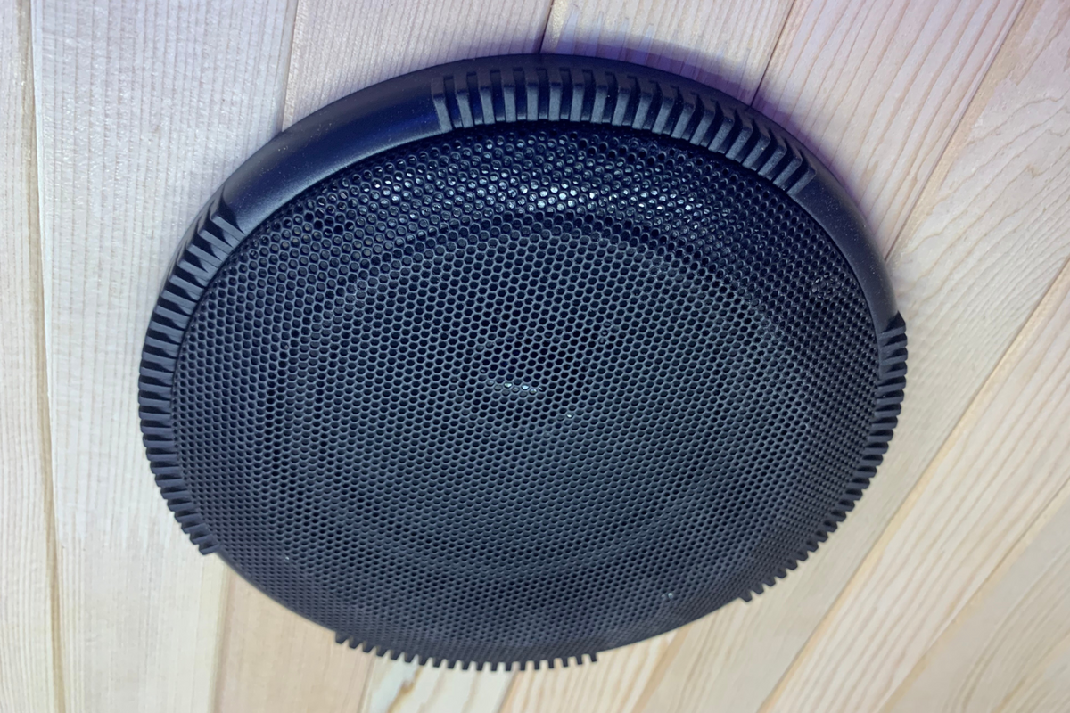 Black speaker grille mounted on a wooden ceiling in a sauna setting.
