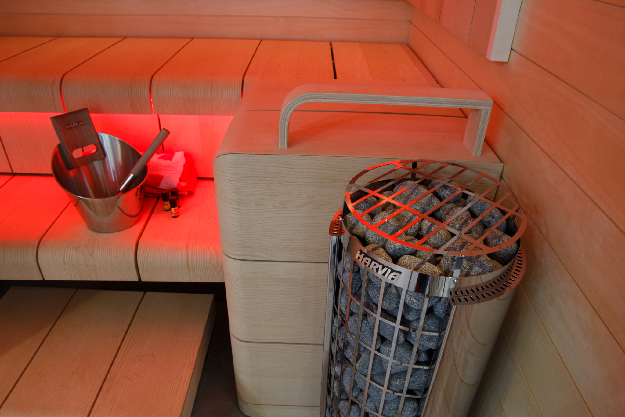 Harvia Cilindro Half PC80E 8kW Electric Sauna Heater in Sauna interior with wooden benches and a metal basket containing stones.