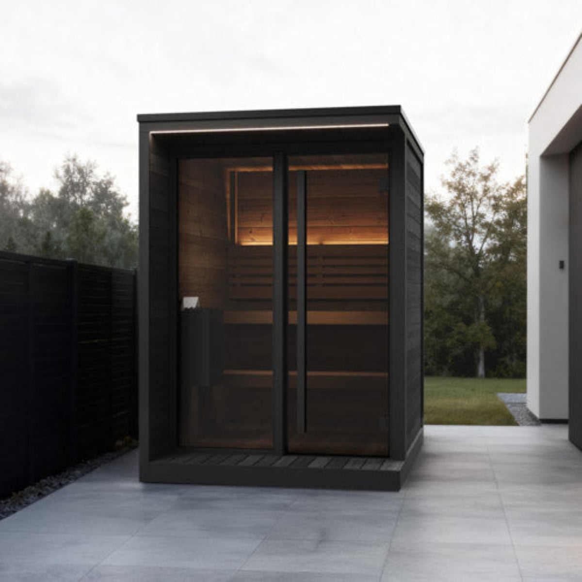 SaunaLife Garden Luxury-Series GL4B with dark wood exterior, large glass doors, and warm interior lighting, situated on a paved patio with trees and a building visible in the background.