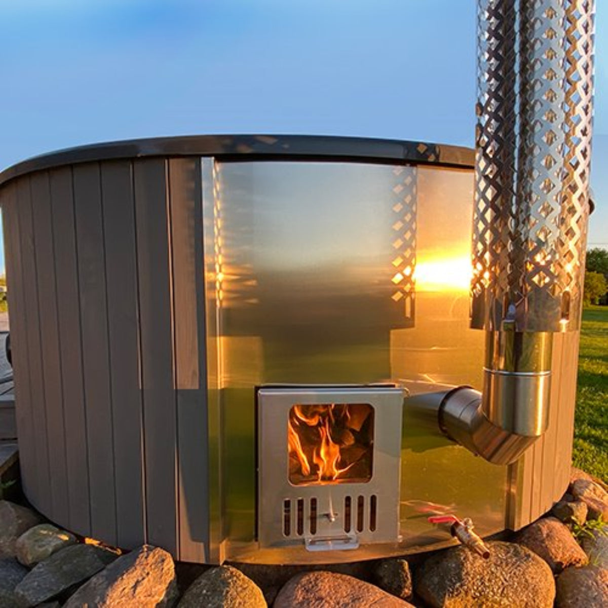 SaunaLife S4N wood-fired hot tub, with flames inside, surrounded by stones and grass with a sunset in the background.