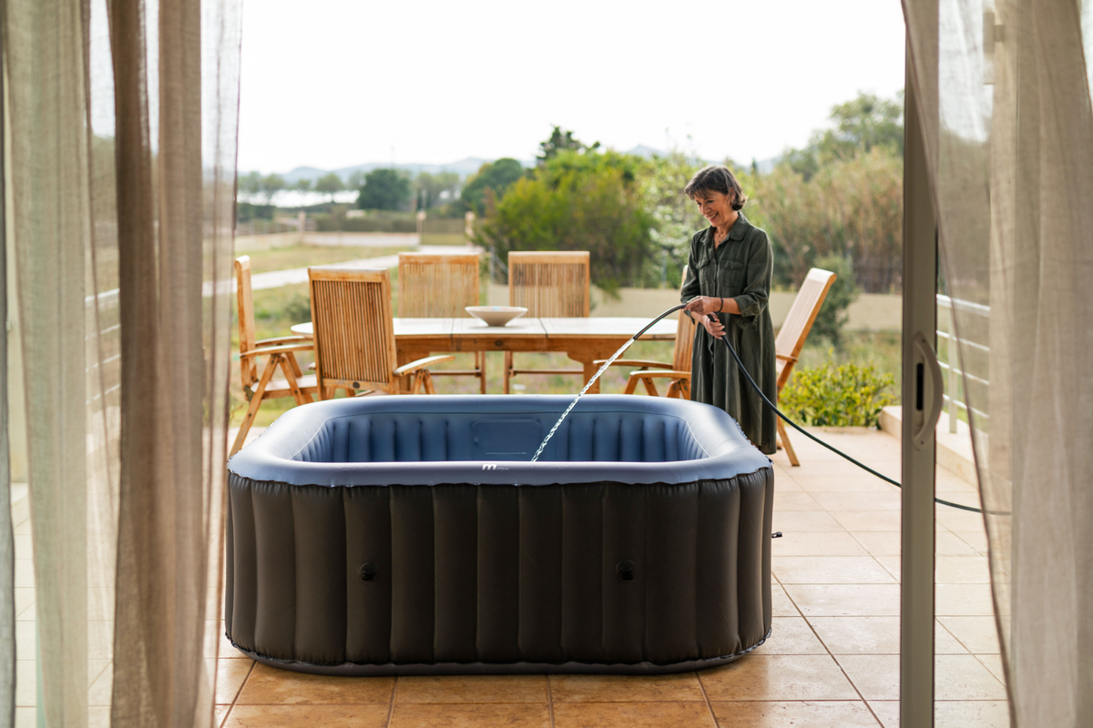 Person filling an inflatable hot tub on a patio with outdoor furniture and greenery in the background.