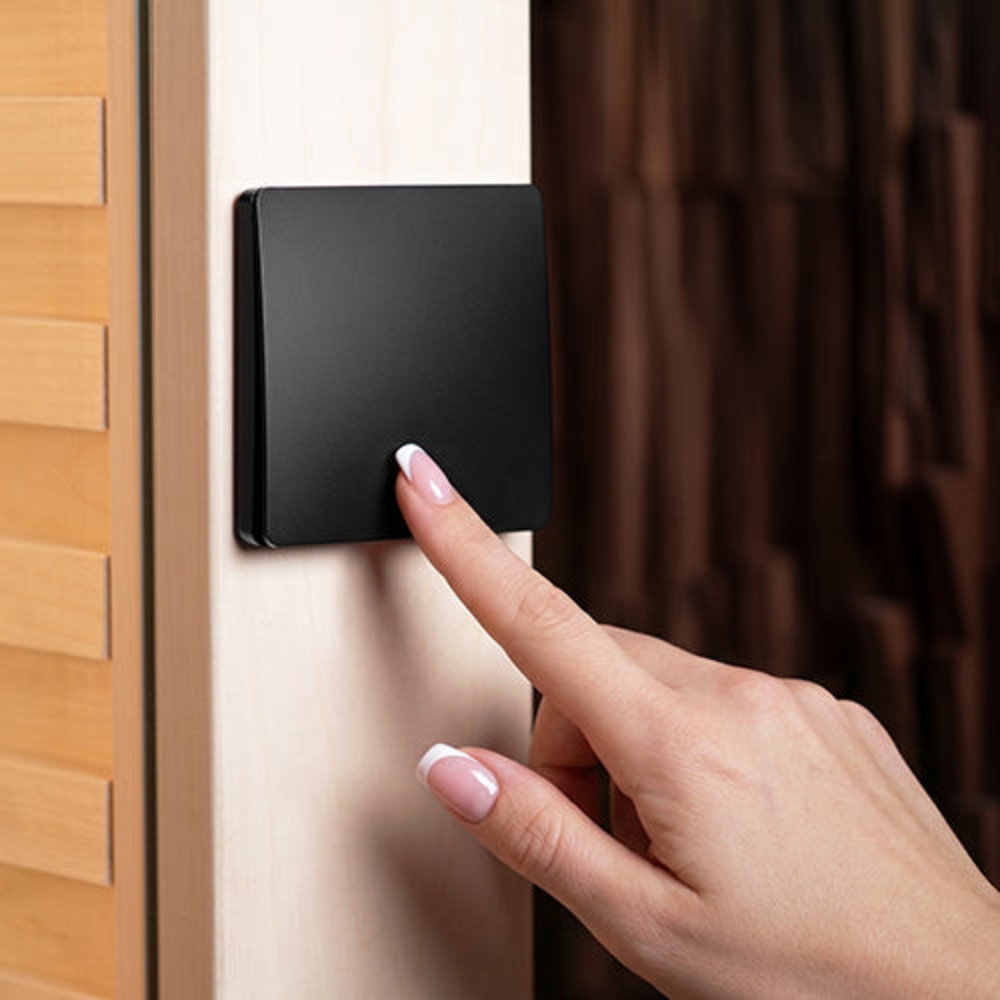 A close-up of a hand pressing a matte black square smart switch on a light wood door panel for Prosaunas led lights