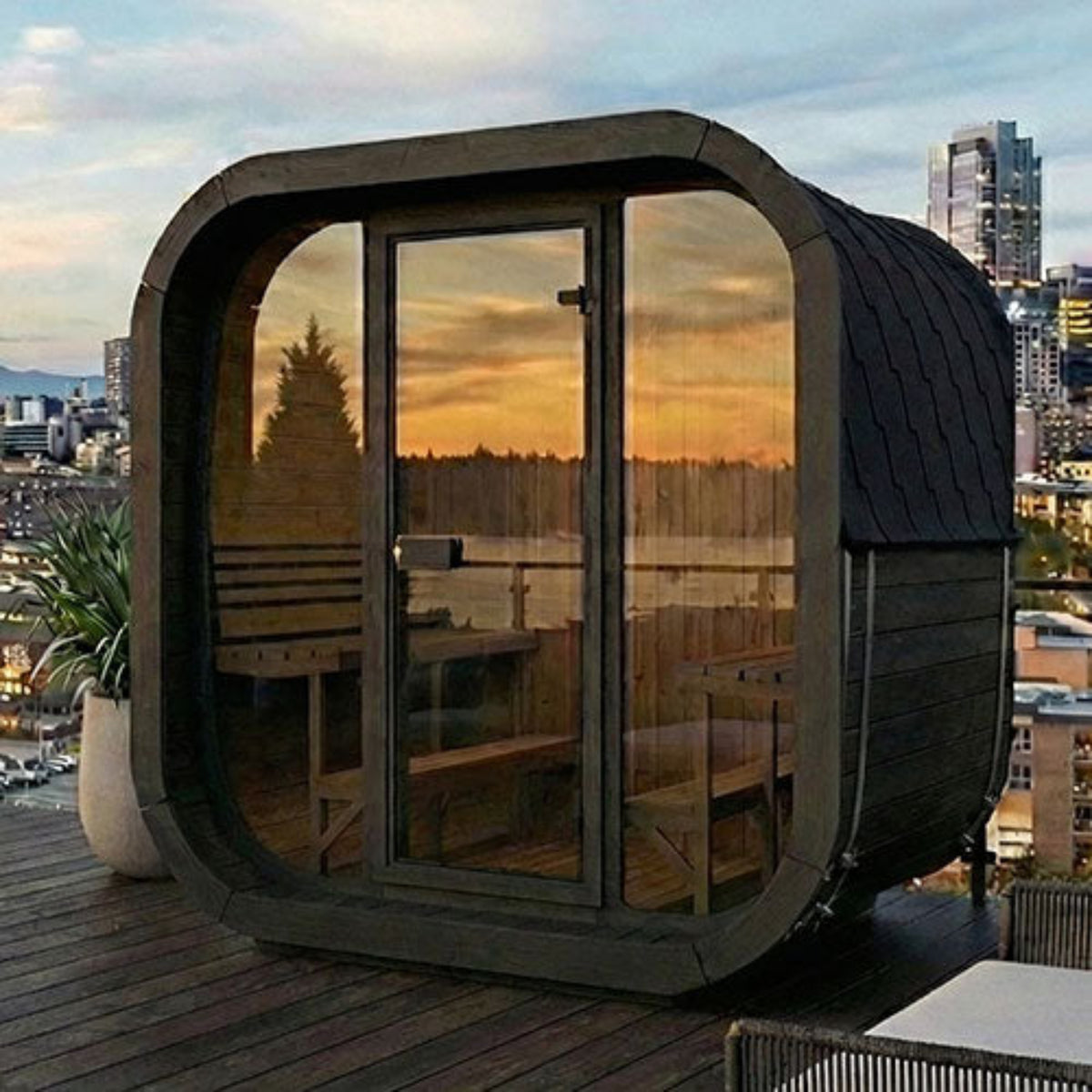 SaunaLife CUBE-Series Model CL7GB Outdoor Cube Sauna with a dark wood exterior, large glass windows, and a curved front design, shown on a rooftop deck with a city skyline and sunset reflections.