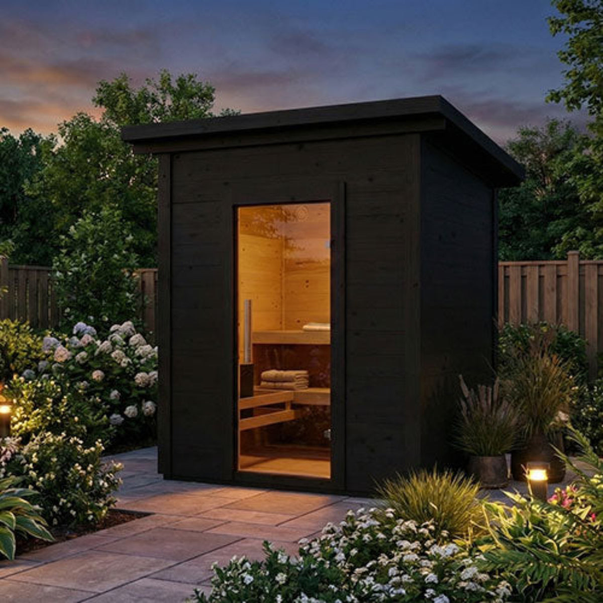 SaunaLife Garden-Series Model G2B Outdoor Garden Sauna with a glass door, illuminated from within, set in a landscaped garden at dusk with flowers, plants, and a fence in the background.