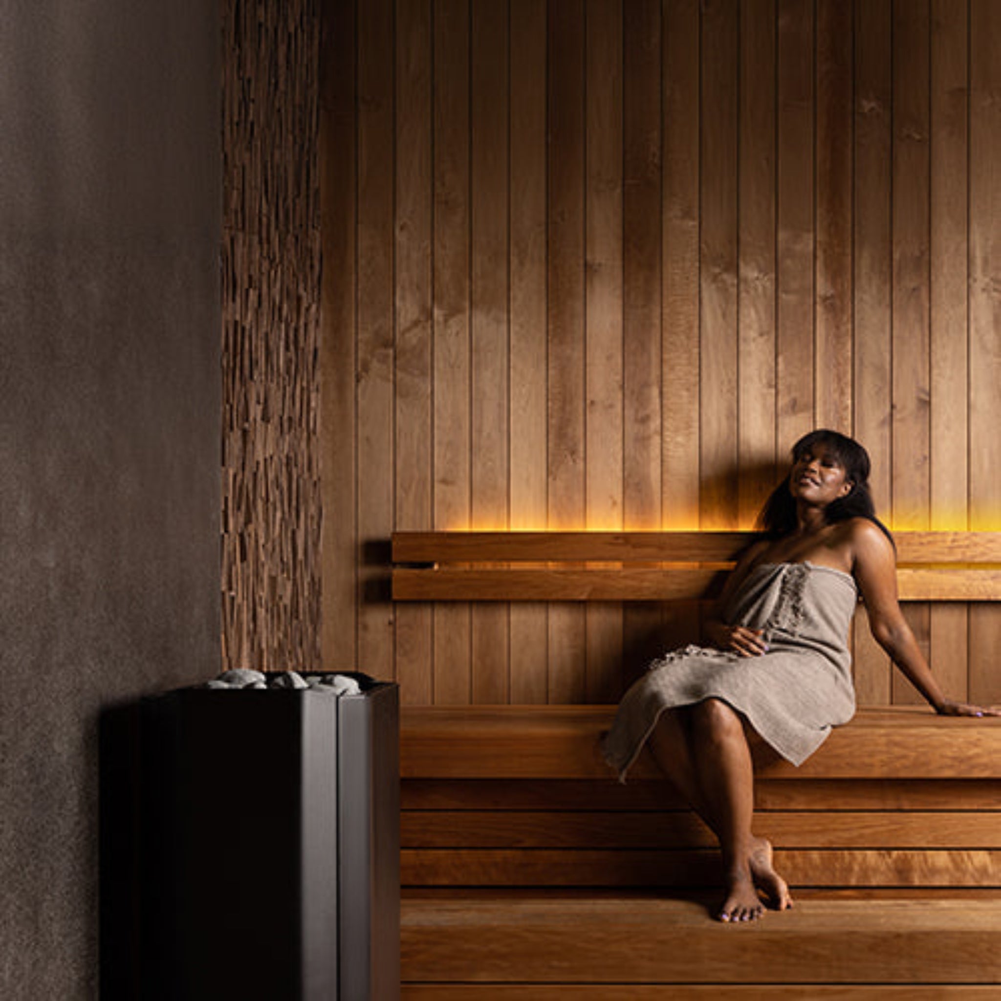 Woman sitting on a wooden bench in a sauna with warm lighting showing a Saunum air perfect sauna heater.